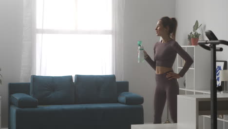 sporty-woman-is-drinking-water-during-break-at-home-workout-relaxing-in-living-room-at-sunny-day-sport-activity-and-healthy-lifestyle